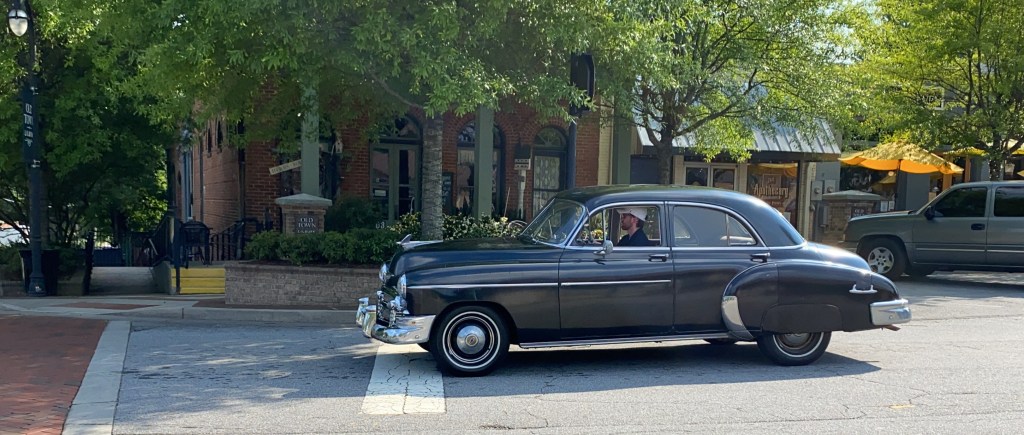 bearded young man in a cap drives a 1950 Chevrolet by brick buildings in downtown Lilburn