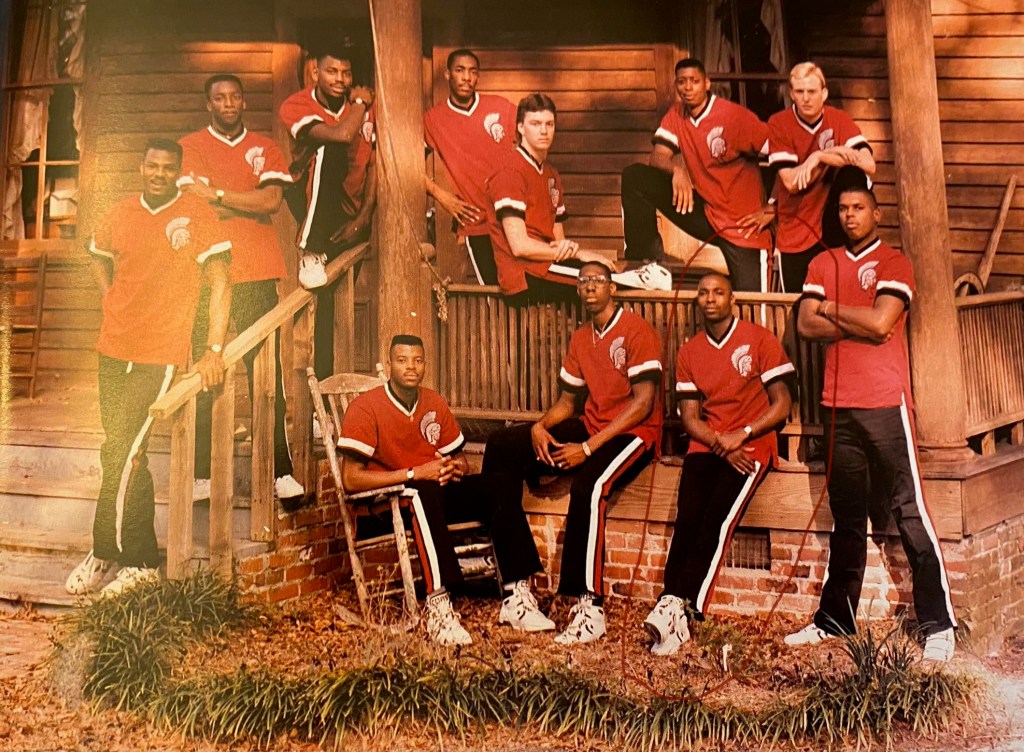 1989 Troy State University Trojan men's basketball team pose for a team photo on the porch of a cabin.