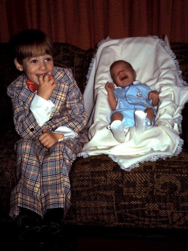 A young boy in a plaid suits sits next to a screaming baby in a baby carrier on a sofa.