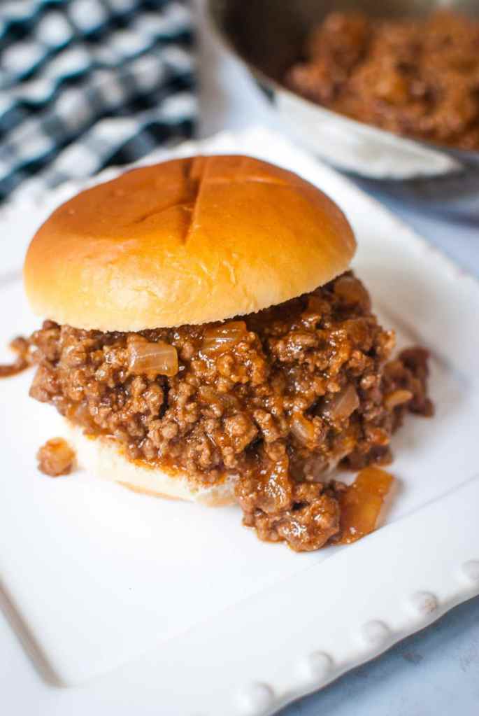 A sloppy joe sandwich on a white rectangular plate with a blue gingham cloth in the background