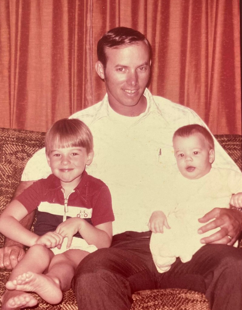 Larry Wallace sitting on a green sofa with his two young sons, Lance and Lee, in his lap.
