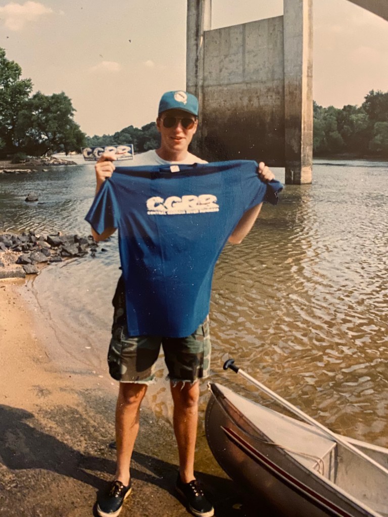 Lance Wallace holds up a Central Georgia River Runners T-shirt beside an aluminum canoe at the shore of the Altamaha River in Lumber City, Georgia, with the Uvalda Bridge in the background.