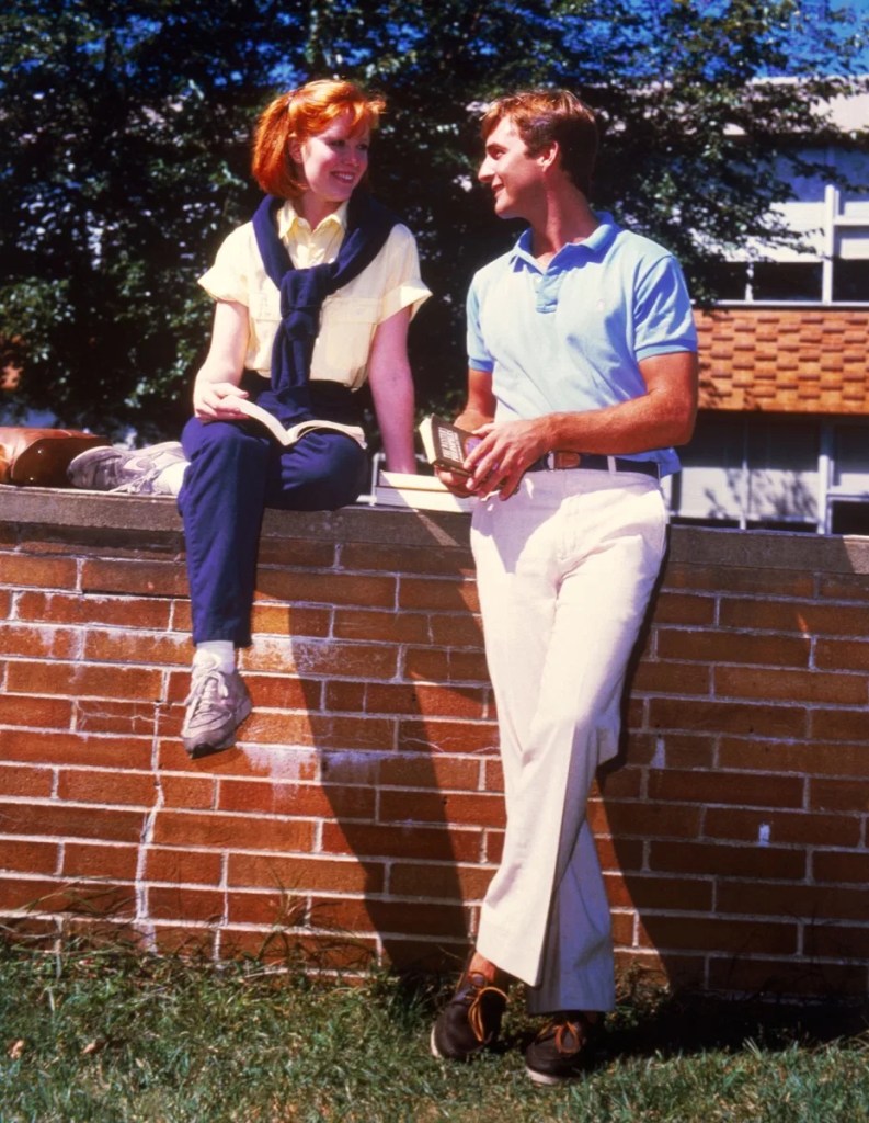 A red-haired, teen-aged girl in blue pants, a yellow shirt with a navy sweater wrapped around her neck and Sperry Top Siders talks with a young man in a light blue polo, white pants and brown boat shoes. They are both holding books. She is sitting on a brick wall in front of a school, and he is leaning against the wall.