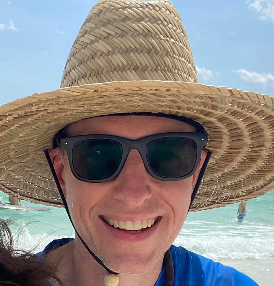 Lance Wallace in straw hat and sun glasses with beach and blue-green waters in the background.