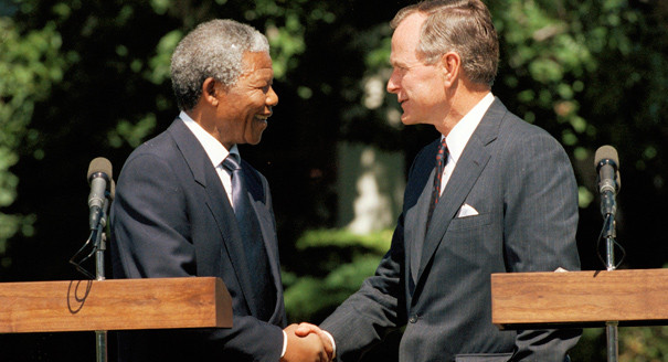 Nelson Mandela, left, shakes hands with U.S. President George H.W. Bush, right, behind two podiums with microphones.