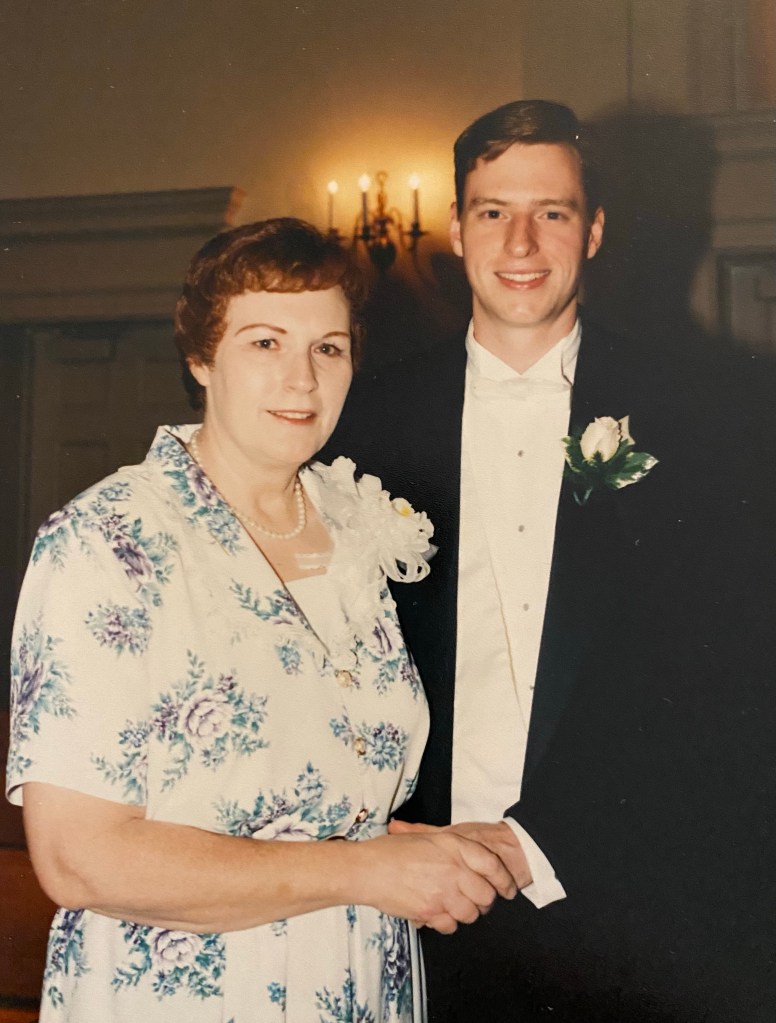 Wearing a tuxedo on his wedding day, Lance Wallace stands with his mother, Sharon Wallace, hold hands in a church with a lit wall sconce in the background of the church sanctuary..