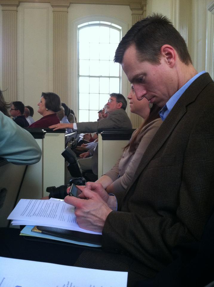 Lance sits on a church pew in a small chapel full of people looking forward. His head bowed, Lance types on a smartphone.
