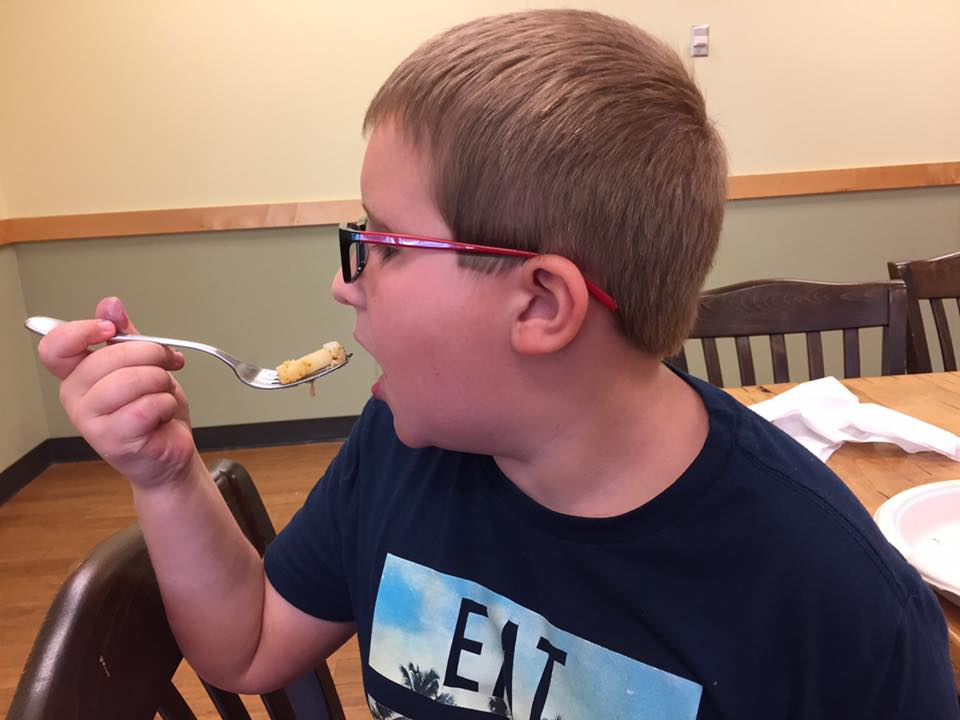 Carlton Wallace in a blue T-shirt holds a fork of french toast ready to eat it with wooden tables and chairs in the background at the Salud Cooking School at Whole Foods in John's Creek, Georgia.