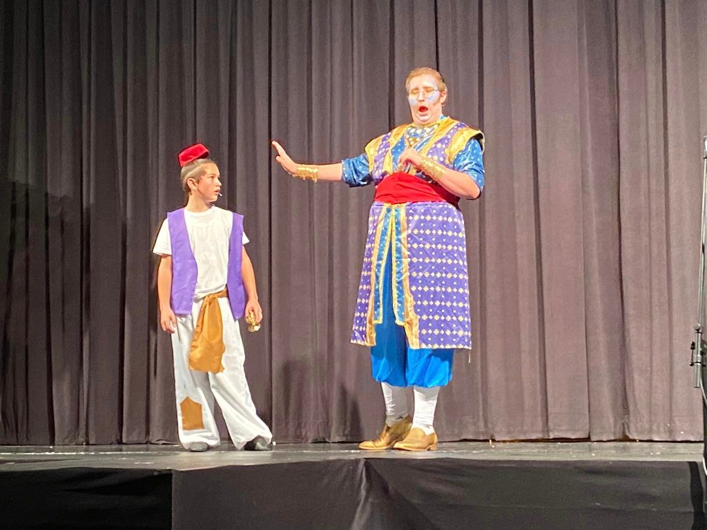 Carlton Wallace dressed in a purple and blue with a blue painted face portrays the genie in a scene with Aladdin on stage in front of a black curtain in a production of the musical Aladdin Jr. this fall.