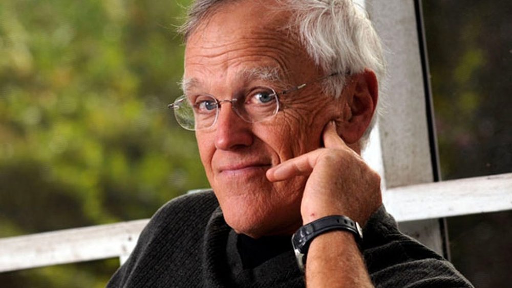 Clyde Edgerton, wearing glasses and a dark sweater, sits on a porch with trees in the background.