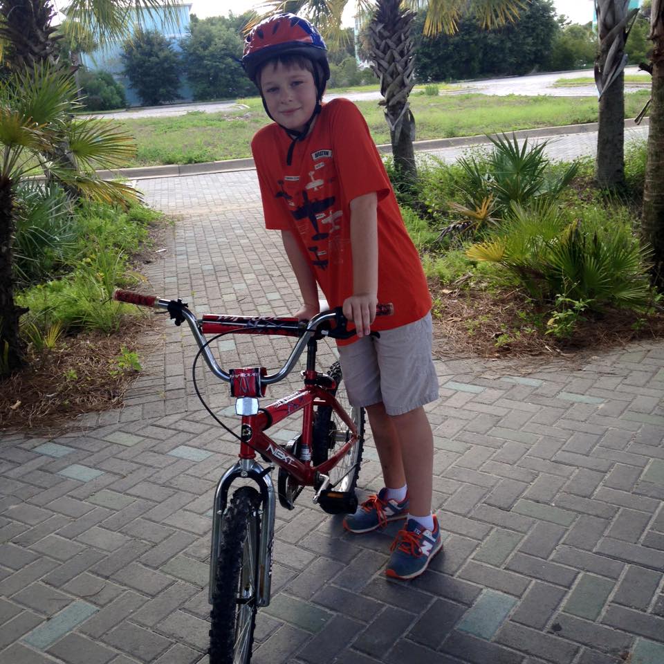 Harris Wallace wearing an orange t-shirt, khaki shorts and a blue and orange bicycle helmet stands with his red bicycle among palm trees and palmetto bushes in Santa Rosa Beach, Florida.