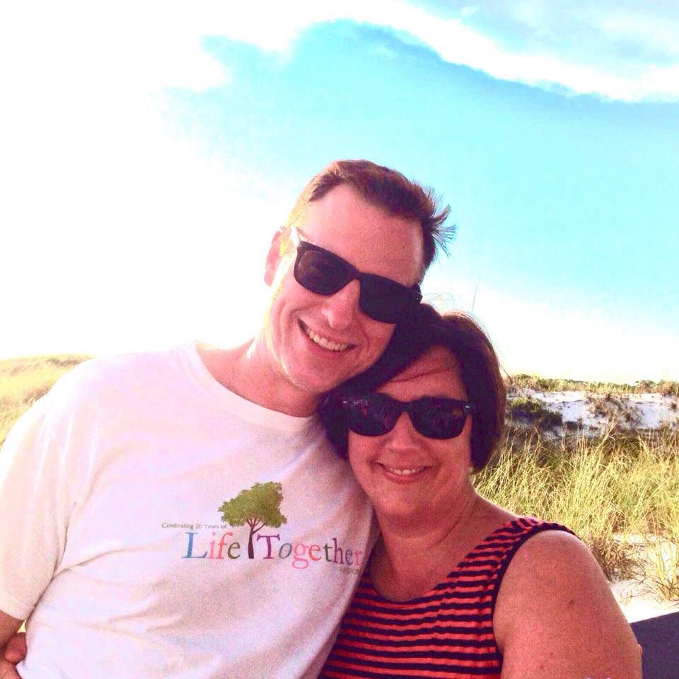 Lance and Carla hug in front of a sand dune at Grayton Beach under a blue sky with white, fluffy clouds.