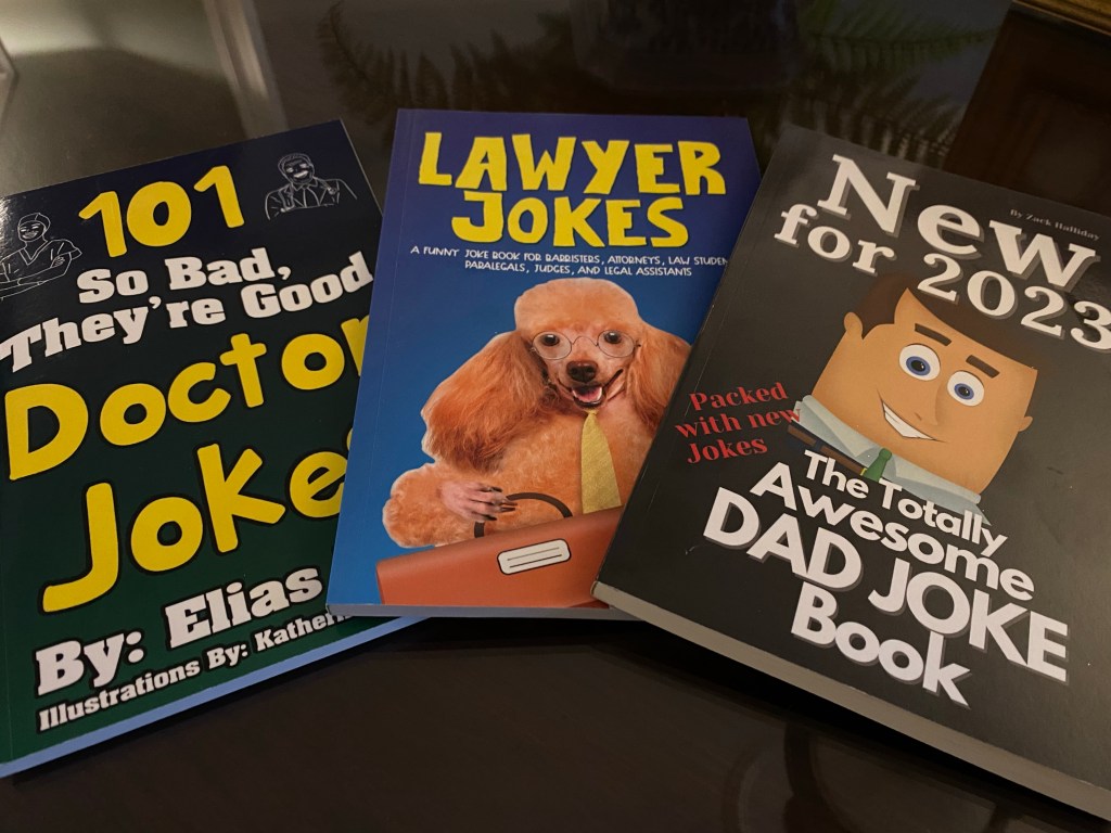 A doctor joke book, lawyer joke book and dad joke book lined up next to each other on a glass table top.