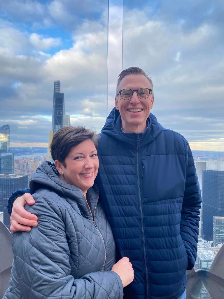 Carla and Lance Wallace embrace on top of Rockefeller Center with the New York City skyline behind them.