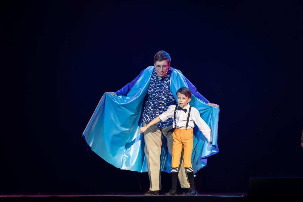 A teenaged boy in a blue cape on stage with a little boy in orange pants and suspenders