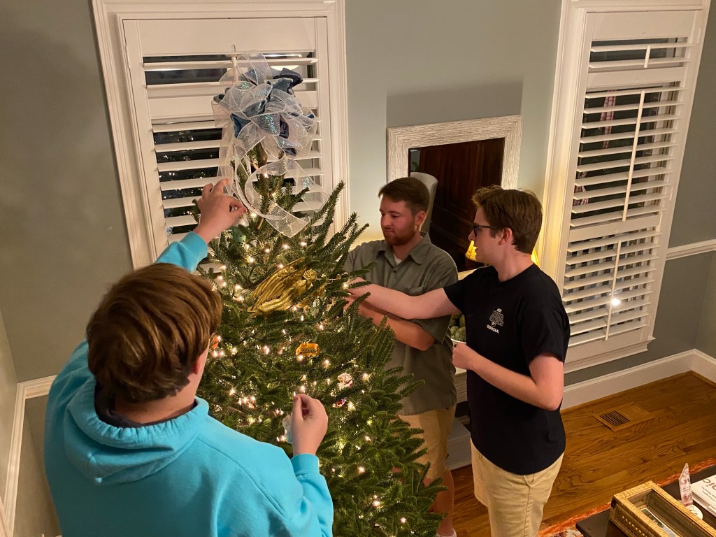 Three young men hang ornaments on a Christmas tree.