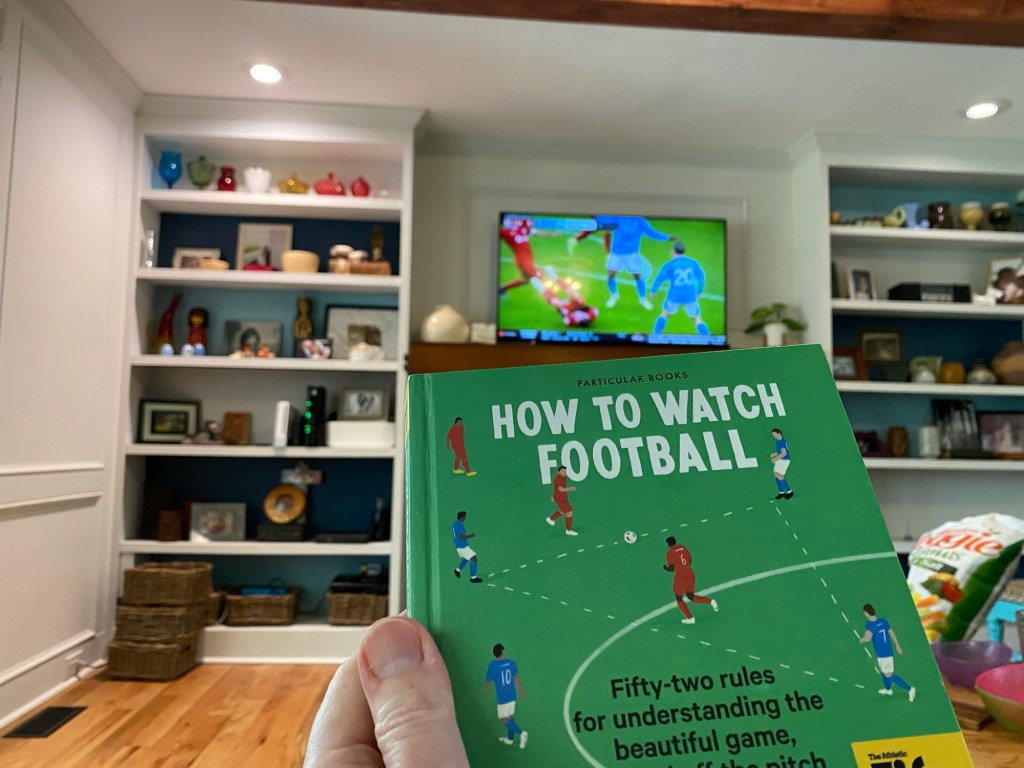a book titled "How to Watch Football" in the foreground with soccer on television in the background