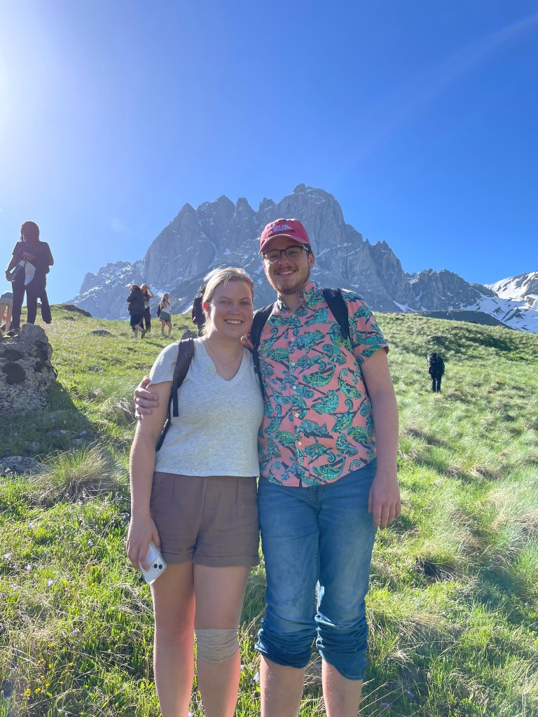 A young woman and young man stand on a grassy slope with a snow-capped mountain in the background