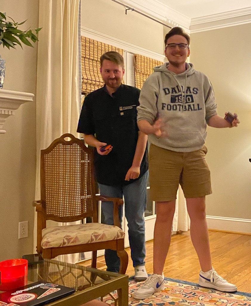 Two young men standing with a young woman sitting on a sofa in a living room