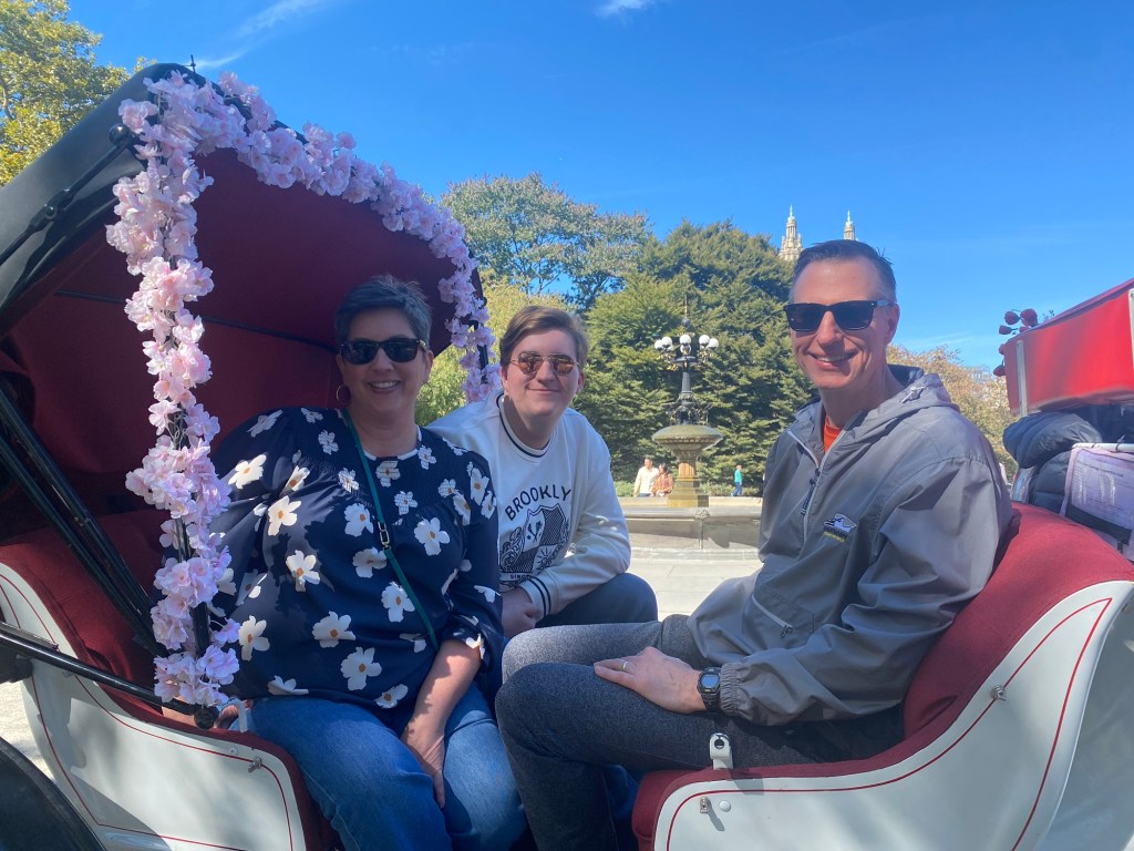 Two men and a woman in a horse-drawn carriage in Central Park.