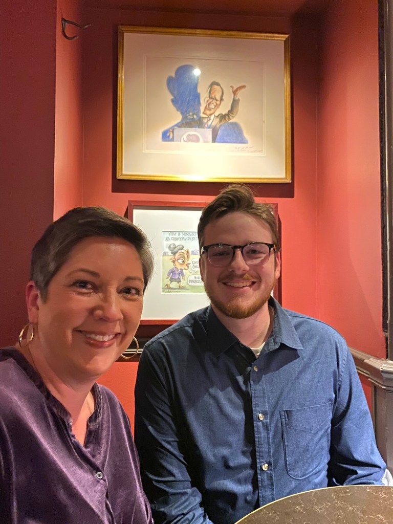A young man and woman at a table with a framed political cartoon of George H.W. Bush on the wall behind them