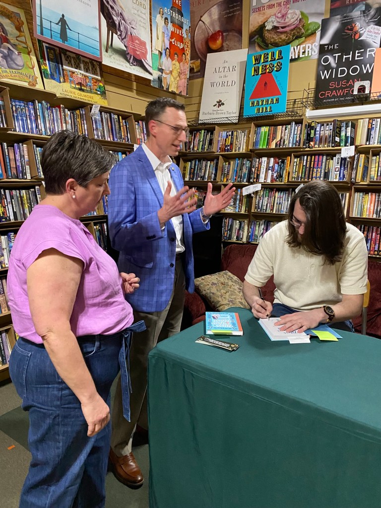 A man talks exaggeratedly with a man signing a book as a woman looks on