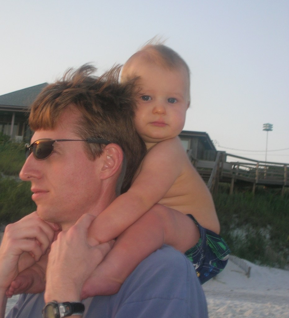 A man in sunglasses with a toddler on his shoulders at the beach