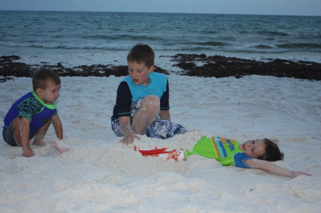 Two boys bury their brother in sand at the beach