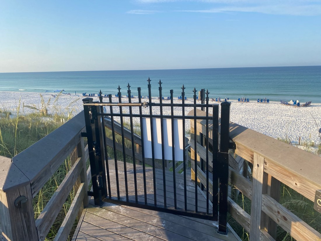 black iron gate secured with a magnet lock on a beach access stair with the white sandy beach and the Gulf in the background