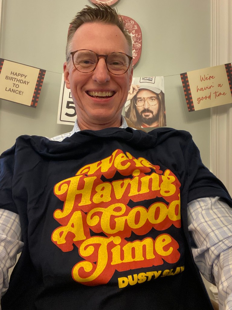 A man with a black T-shirt with the words "We're Having a Good Time" and "Dusty Slay" on it.