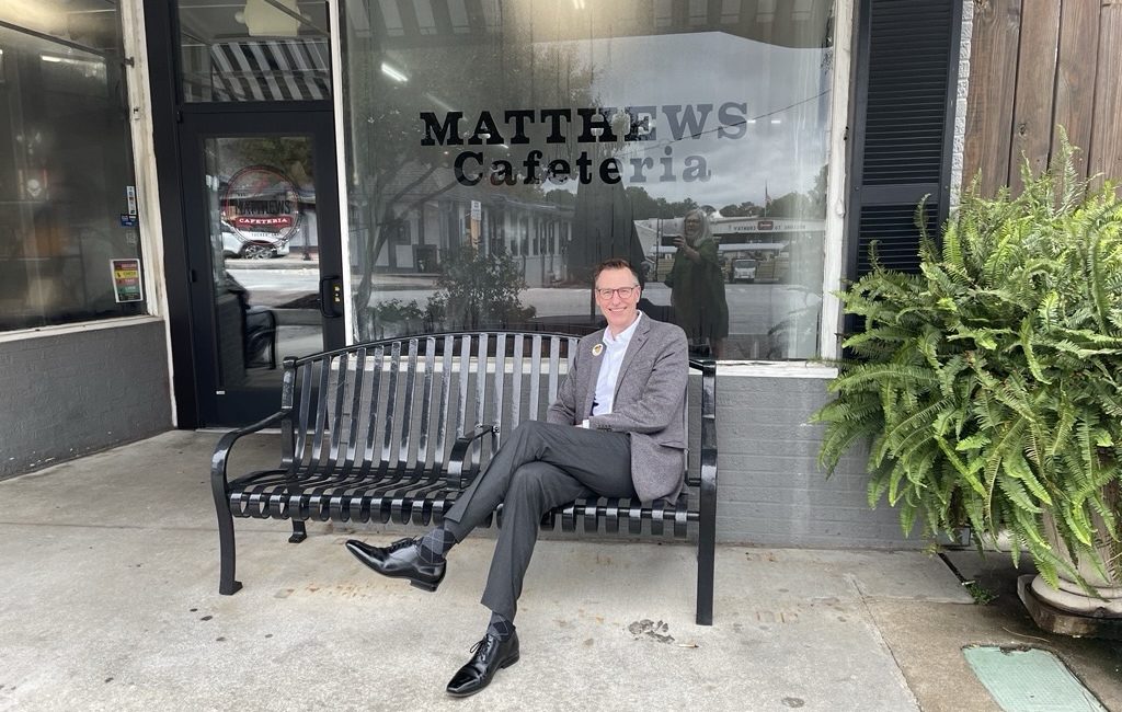 A man on a bench in front of a window that reads "Matthews Cafeteria"