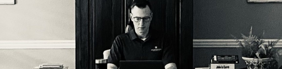 A black and white photo of Lance Wallace sitting at his desk typing on his laptop.