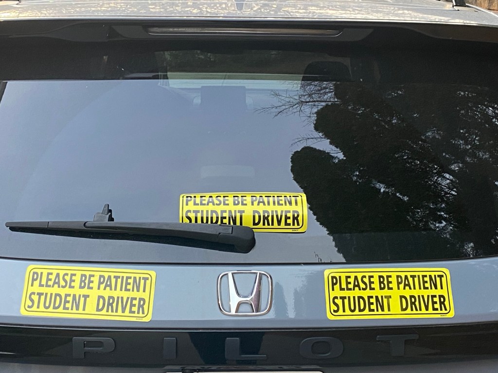 Back of a pearl gray Honda Pilot with three yellow magnets that say "Please be patient student driver"
