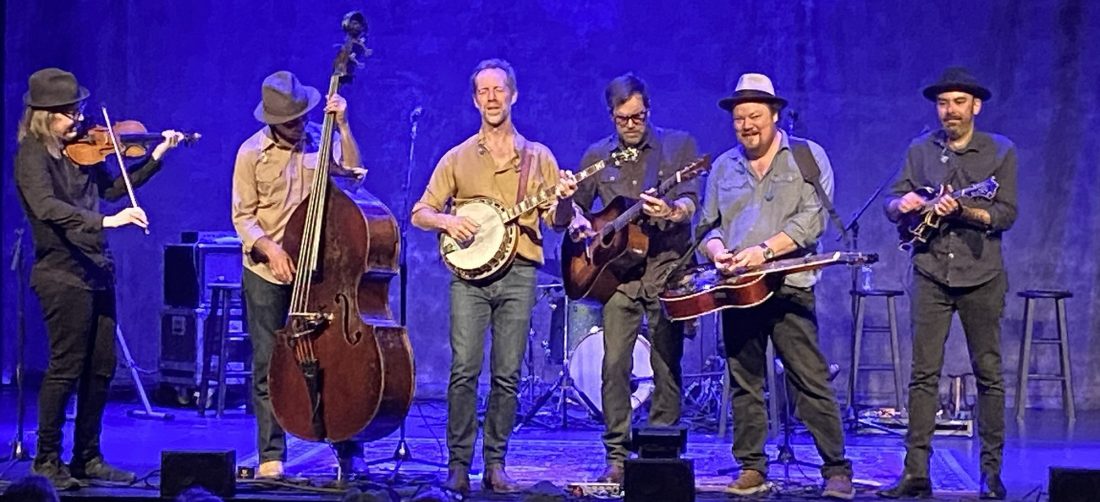 Six members of a bluegrass band on a stage lit with blue lights playing their instruments.