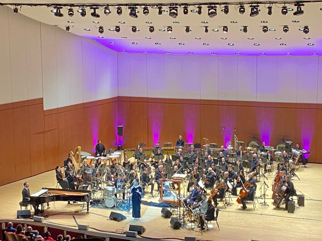 A woman in a black velvet dress sings on a stage surrounded by an orchestra