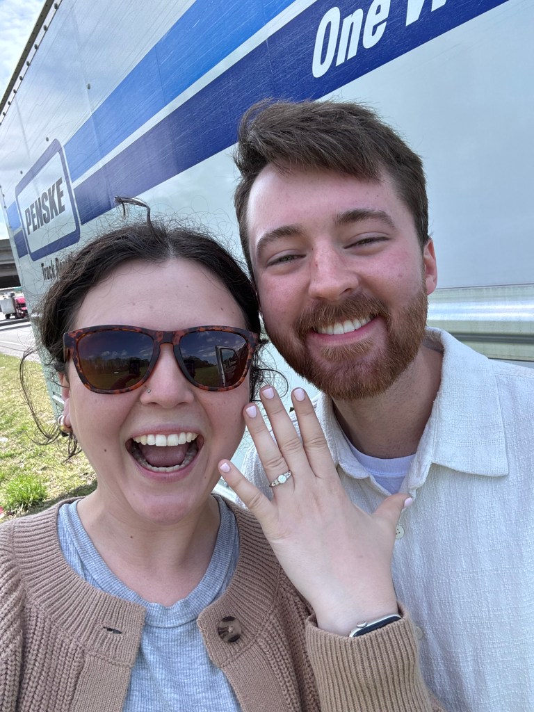 A young woman in sunglassed in a brown sweater displays her engagement ring with a young bearded man looking on.