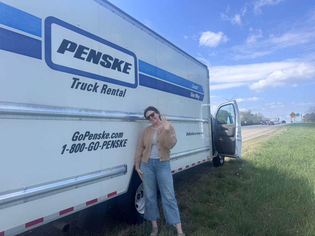 A young woman in a brown sweater holds up her hand displaying an engagement ring stands beside a white Penske truck on the side of a highway.