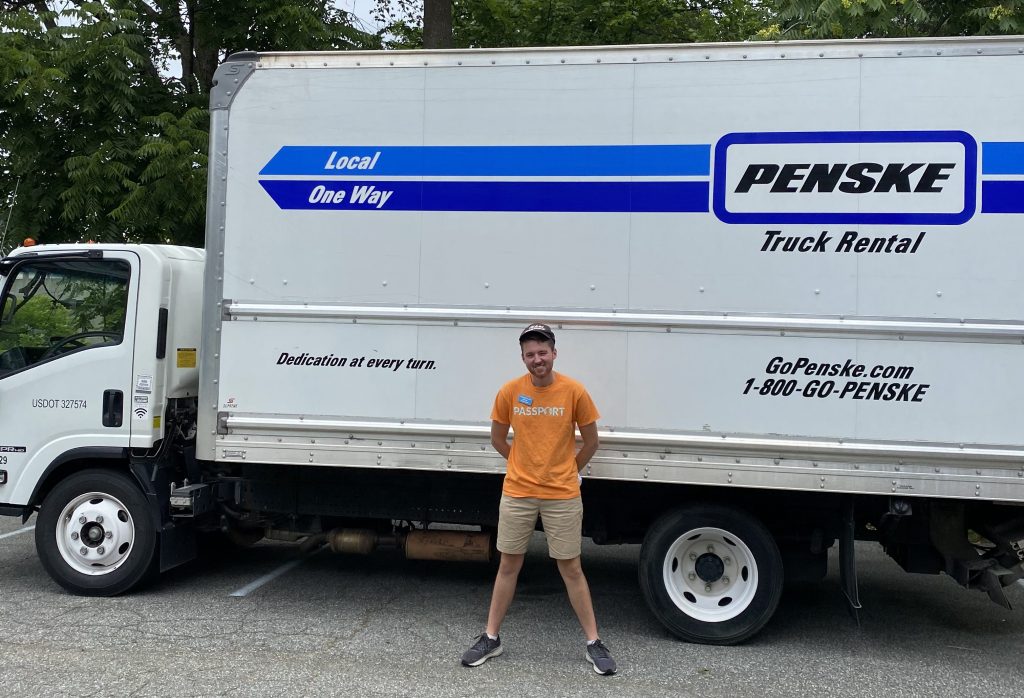 A young man in an orange T-shirt standing in front of a white rented Penske truck.