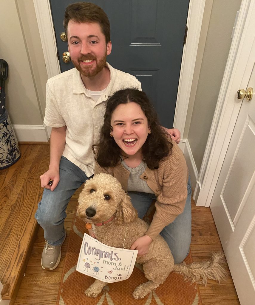 A happy couple kneeling with their golden doodle wearing a sign that says "Congrats Mom and Dad."