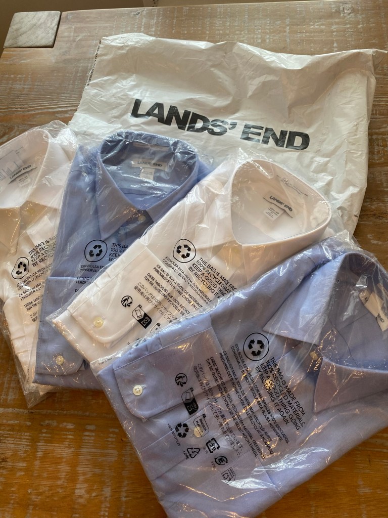 Two white dress shirts and two blue shirts wrapped in plastic on top of a Land's End package on a table.
