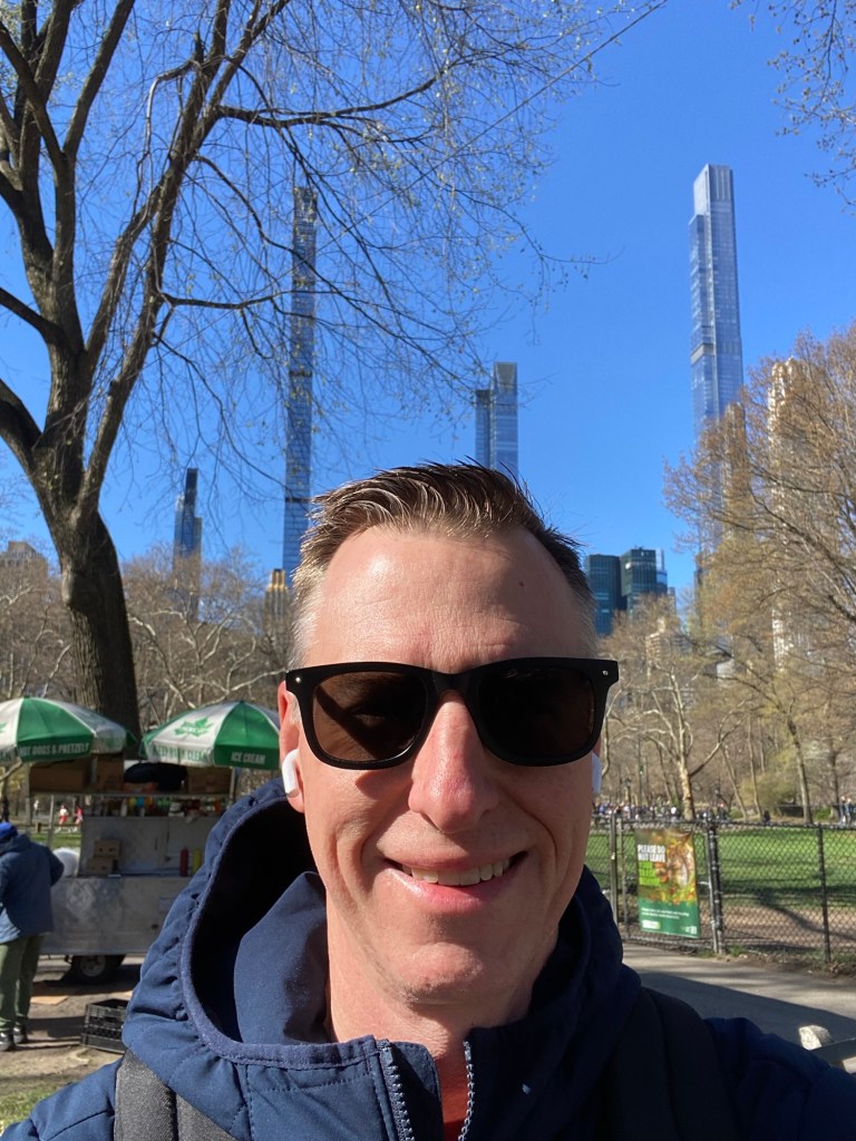 Selfie of a man in sunglasses in Central Park with tall buildings in the background.
