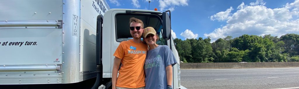 A young man and woman stand outside the cab of a white rented Penske truck on the side of a highway.