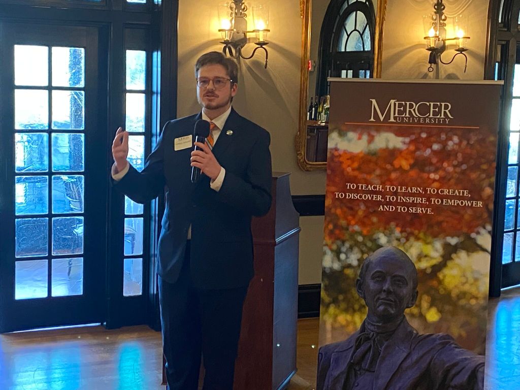 A young man in a suit and tie gives a speech with a microphone in front of a Mercer University banner