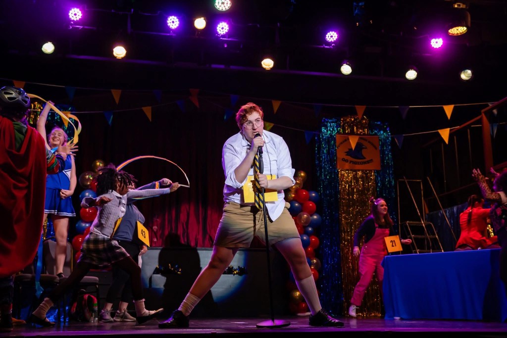 A teenager in a disheveled white shirt, striped necktie and khaki shorts sings into a microphone.