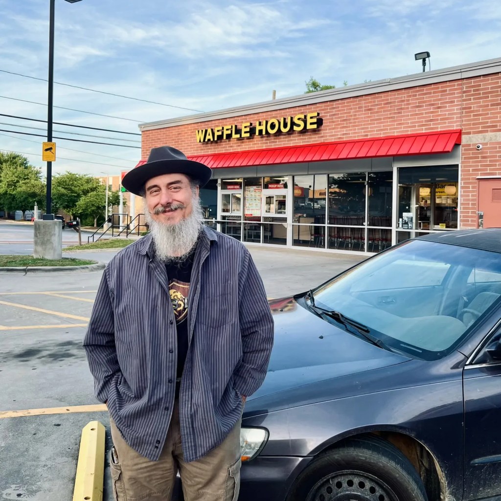 A man with a twirled mustache and beard wearing a hat outside of a Waffle House