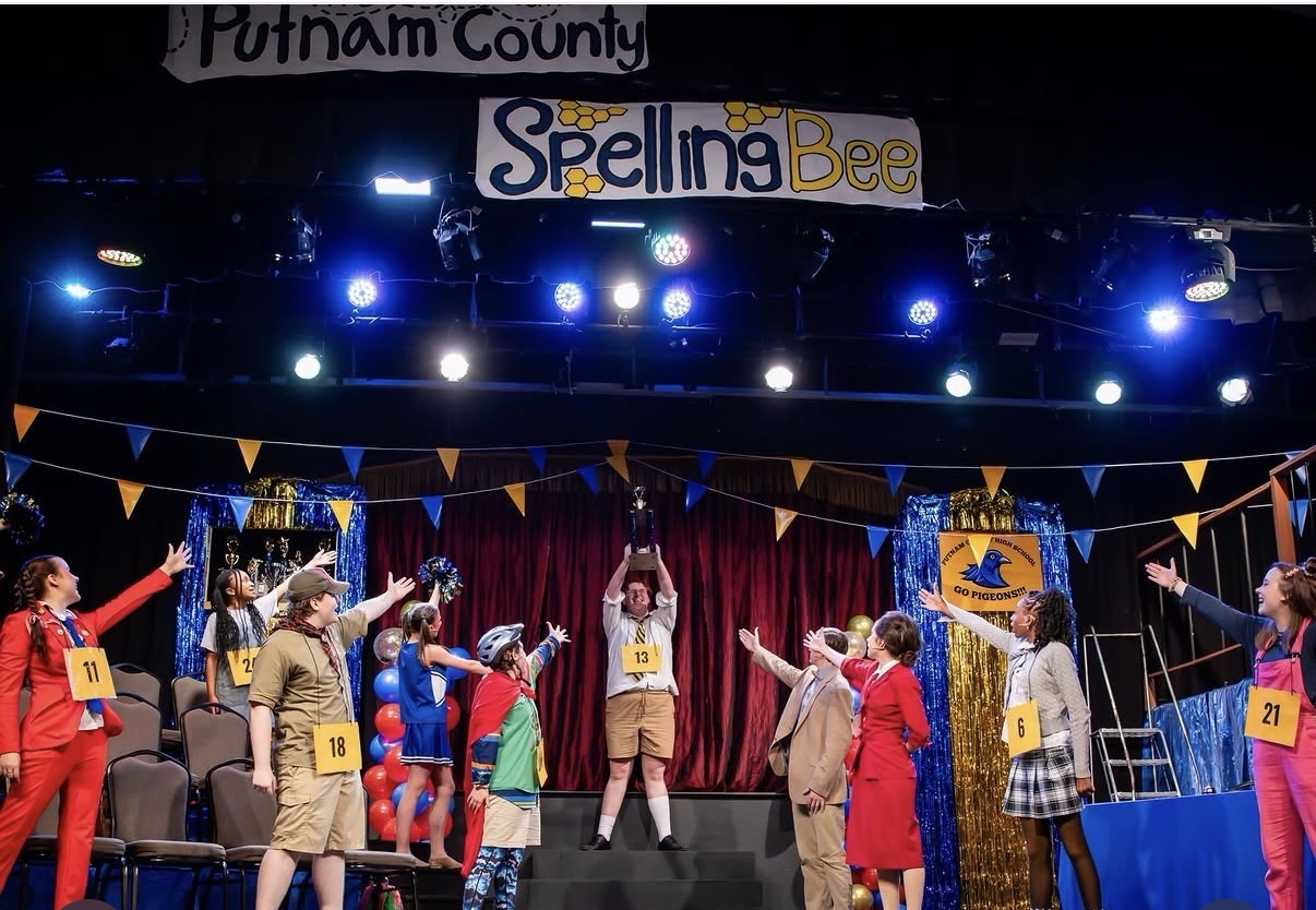 A teenager holds a trophy over his head on a stage under a banner that reads "Putnam County Spelling Bee"