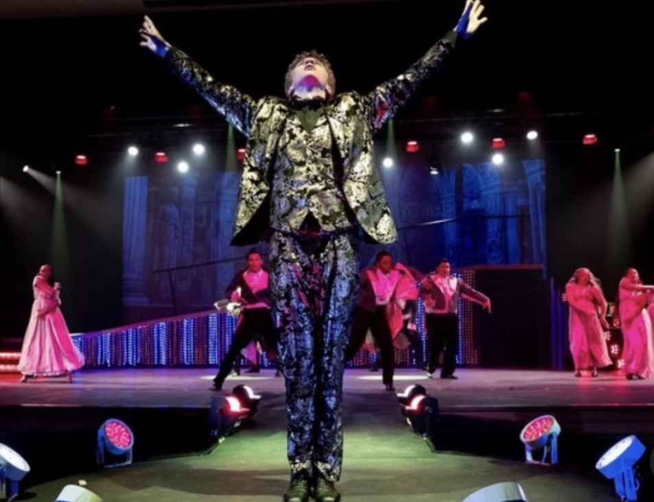 Prince Charming with his arms raised on stage as teenagers dance in the background