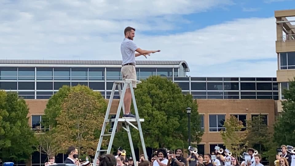A band drum major at the top of a metal ladder with the band below him.