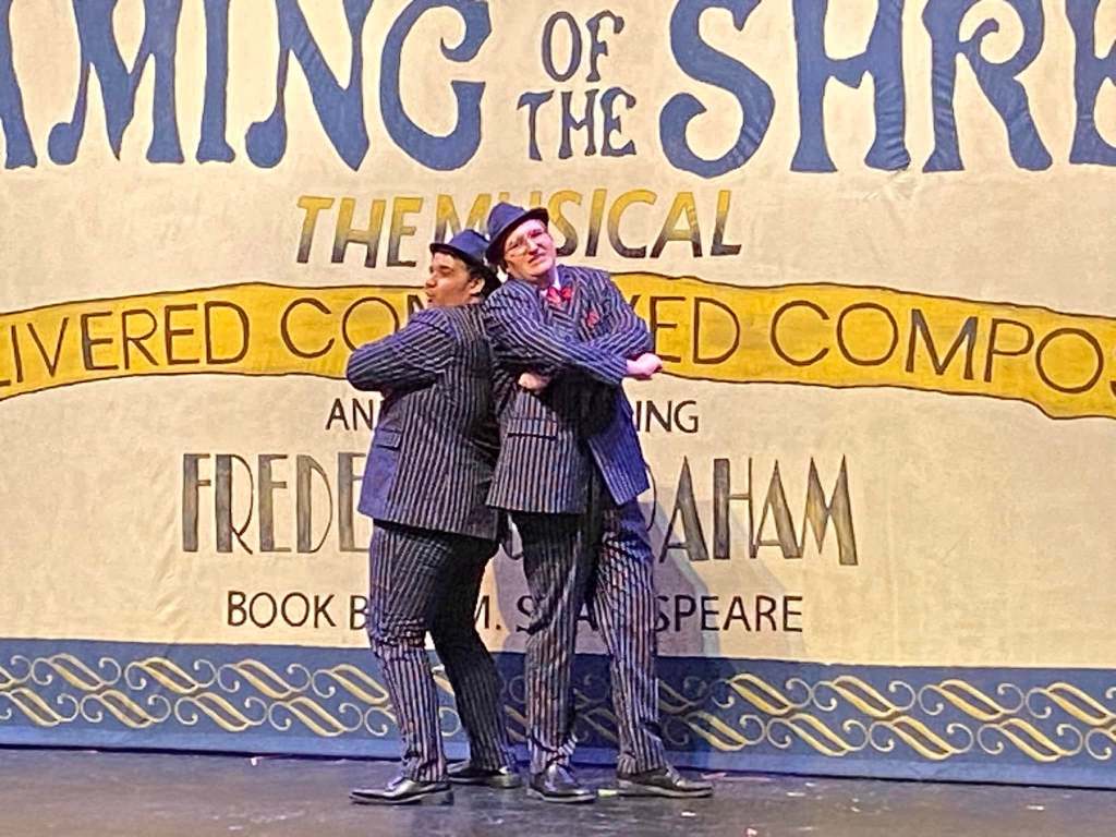 Two gangsters in navy pinstriped suits and blue fedoras stand with arms crossed on stage in front of a painted curtain.