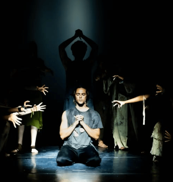 An man kneels with his hands folded in prayer as a man holds a knife over his head